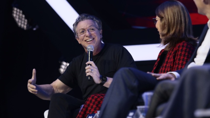 Boninho sorridente, ao lado de Daniela Beyruti e executivo na Disney, em trecho da coletiva do The Voice Brasil