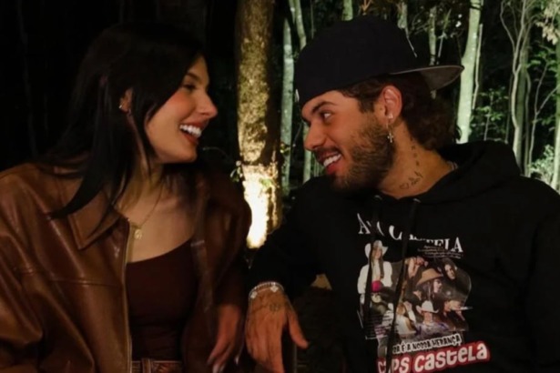 Os cantores Ana Castela (cabelos longos e escuros, jaqueta de couro marrom e top marrom) e Zé Felipe (boné preto e blusa de moletom preta estampada) sorriem um para o outro em um ambiente externo à noite, com luzes ao fundo.