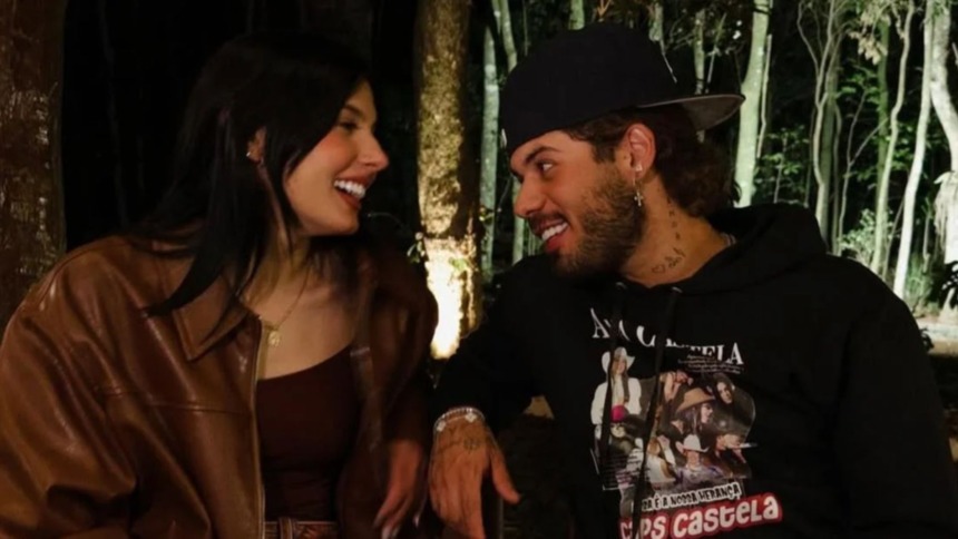 Os cantores Ana Castela (cabelos longos e escuros, jaqueta de couro marrom e top marrom) e Zé Felipe (boné preto e blusa de moletom preta estampada) sorriem um para o outro em um ambiente externo à noite, com luzes ao fundo.