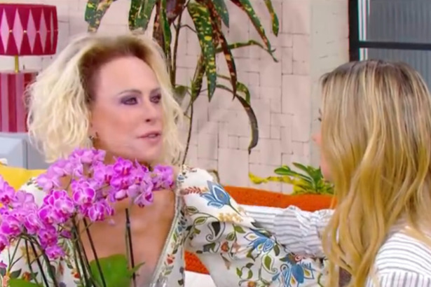 Ana Maria Braga (cabelos loiros, maquiagem pesada) conversa com Carolina Dieckmann (cabelos loiros, de costas) no sofá amarelo do Mais Você, segurando um arranjo de orquídeas roxas.
