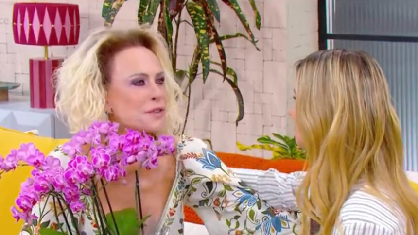 Ana Maria Braga (cabelos loiros, maquiagem pesada) conversa com Carolina Dieckmann (cabelos loiros, de costas) no sofá amarelo do Mais Você, segurando um arranjo de orquídeas roxas.