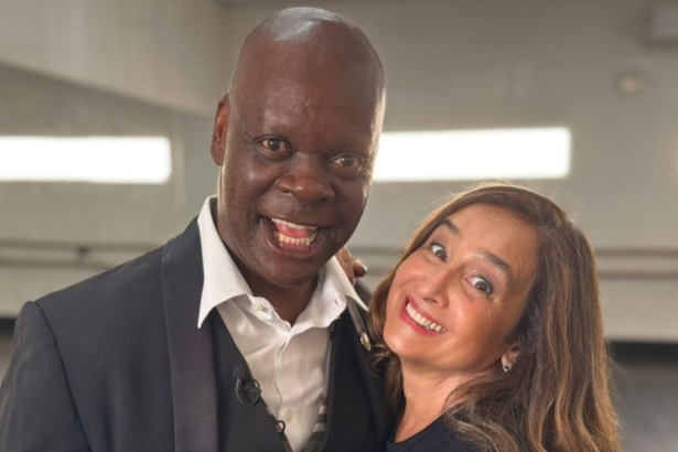 O ator Sebastian, de terno preto, e a jornalista Graziela Azevedo sorriem juntos e de forma animada nos bastidores do Globo Repórter.