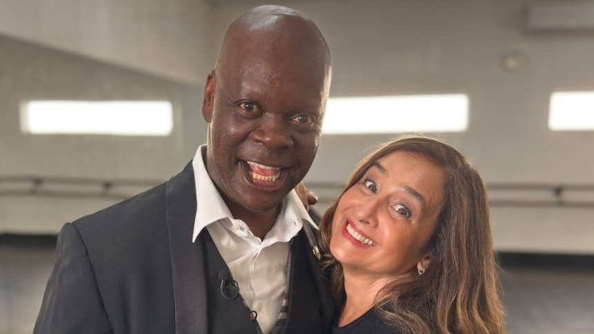 O ator Sebastian, de terno preto, e a jornalista Graziela Azevedo sorriem juntos e de forma animada nos bastidores do Globo Repórter.