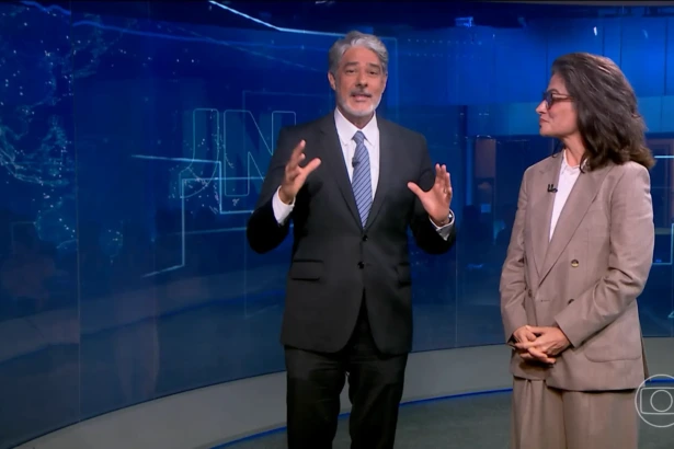 William Bonner ao lado de Renata Vasconcellos em pé no cenário do Jornal Nacional. Ele levanta as mãos para convidar Cesar Tralli