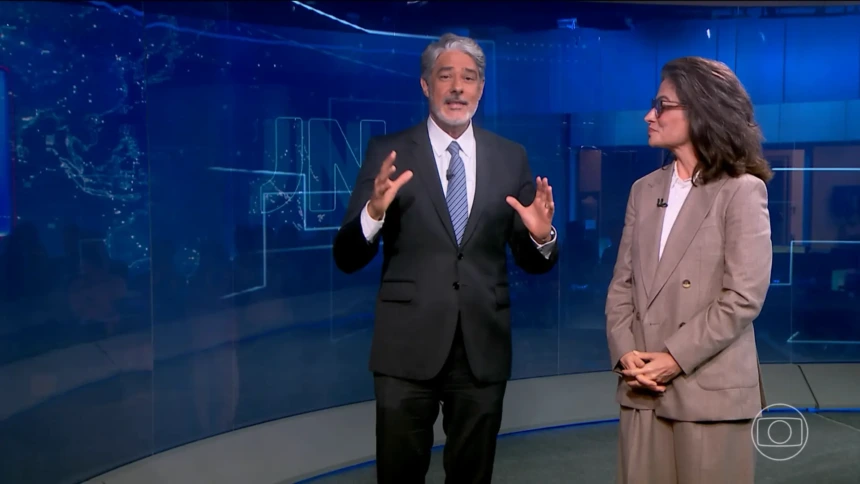 William Bonner ao lado de Renata Vasconcellos em pé no cenário do Jornal Nacional. Ele levanta as mãos para convidar Cesar Tralli
