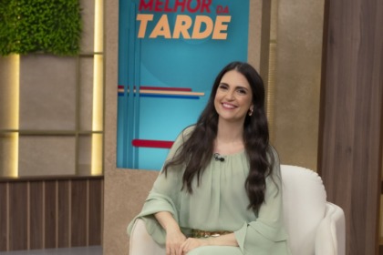 Chris Flores no cenário do Melhor da Tarde. Ela veste um vestido verde e está sentada em uma poltrona