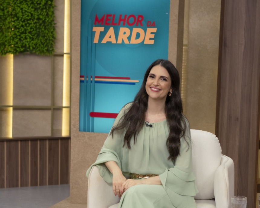Chris Flores no cenário do Melhor da Tarde. Ela veste um vestido verde e está sentada em uma poltrona