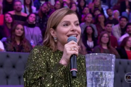 A atriz Debora Bloch sorri, falando em um microfone no Domingão, falando sobre Vale Tudo. Ela usa um vestido verde de lantejoulas e brincos grandes.