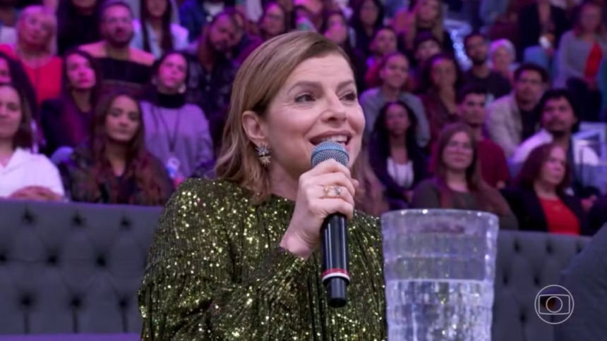 A atriz Debora Bloch sorri, falando em um microfone no Domingão, falando sobre Vale Tudo. Ela usa um vestido verde de lantejoulas e brincos grandes.