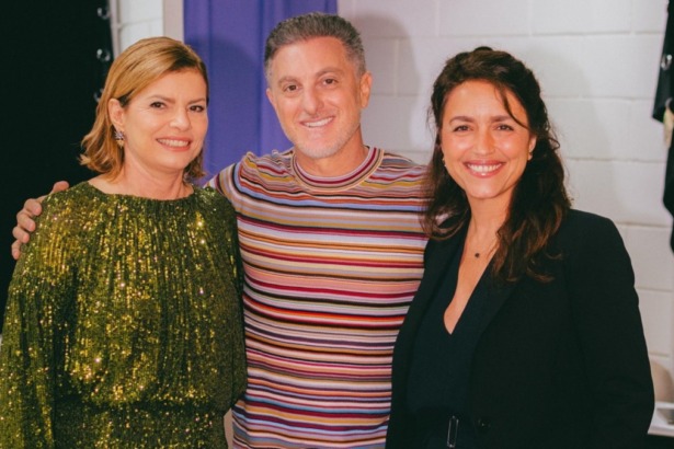 A atriz Debora Bloch (vestido verde de lantejoulas), Luciano Huck (camiseta listrada) e a autora Manuela Dias (blazer preto) sorriem juntos nos bastidores do Domingão
