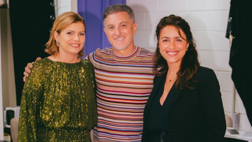 A atriz Debora Bloch (vestido verde de lantejoulas), Luciano Huck (camiseta listrada) e a autora Manuela Dias (blazer preto) sorriem juntos nos bastidores do Domingão