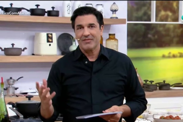 O apresentador e chef Edu Guedes em pé no estúdio de seu programa, "Fica com a gente". Ele veste camisa preta e gesticula em frente a uma cozinha.