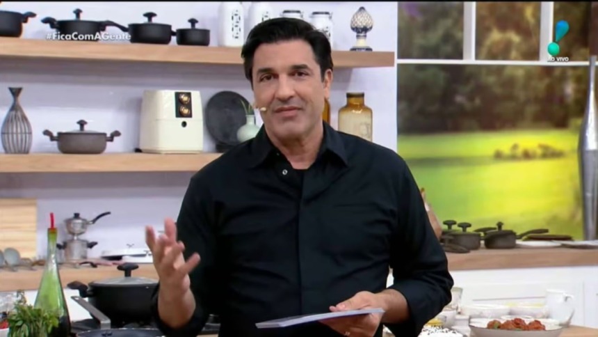 O apresentador e chef Edu Guedes em pé no estúdio de seu programa, "Fica com a gente". Ele veste camisa preta e gesticula em frente a uma cozinha.