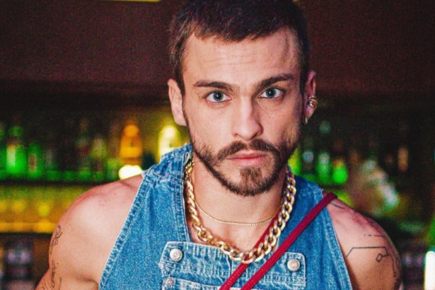 O ator Felipe Selau faz um close-up, olhando intensamente para a câmera. Ele tem barba e usa um macacão jeans com correntes grossas douradas no pescoço.