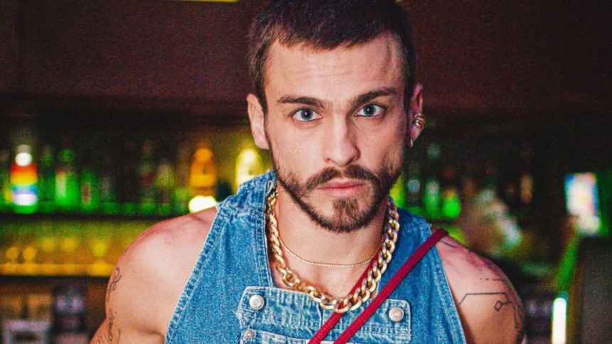 O ator Felipe Selau faz um close-up, olhando intensamente para a câmera. Ele tem barba e usa um macacão jeans com correntes grossas douradas no pescoço.