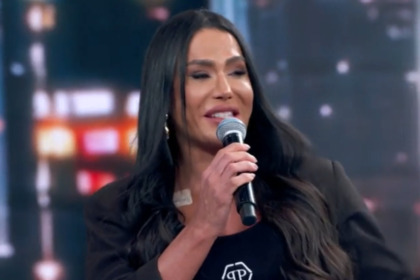 Gracyanne Barbosa, de blazer e top preto, fala em um microfone no palco do Domingão. Ela tem cabelos longos e escuros e uma atadura clara no peito.