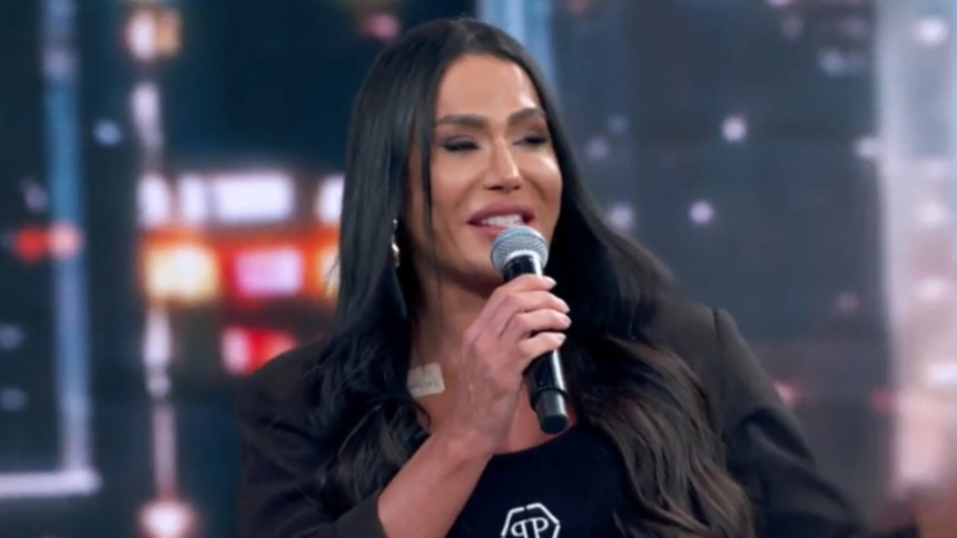 Gracyanne Barbosa, de blazer e top preto, fala em um microfone no palco do Domingão. Ela tem cabelos longos e escuros e uma atadura clara no peito.