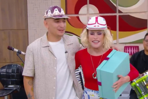 cantor João Gomes e a apresentadora Ana Maria Braga sorriem e se abraçam no cenário do Mais Você, ambos usando bonés temáticos. Ana Maria segura uma caixa azul turquesa.