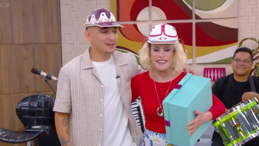 cantor João Gomes e a apresentadora Ana Maria Braga sorriem e se abraçam no cenário do Mais Você, ambos usando bonés temáticos. Ana Maria segura uma caixa azul turquesa.