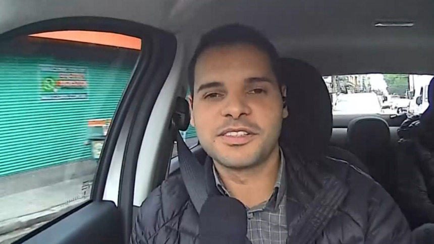 O repórter João Netto em entrada ao vivo no Bom Dia SP. Ele está dentro de um carro, de jaqueta, falando no microfone.
