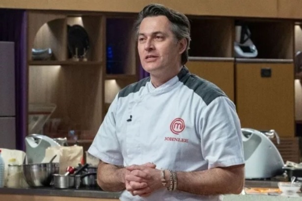 Johnlee está na cozinha, com dólmã branco e preto, e as mãos entrelaçadas. O fundo mostra prateleiras e utensílios de cozinha do MasterChef Confeitaria