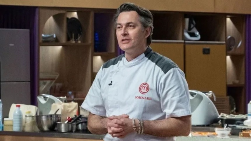 Johnlee está na cozinha, com dólmã branco e preto, e as mãos entrelaçadas. O fundo mostra prateleiras e utensílios de cozinha do MasterChef Confeitaria