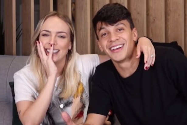 Os influenciadores e apresentadores Júlio Cocielo (camiseta preta) e Tata Estaniecki (cabelo loiro, camiseta branca) sorriem e se abraçam em um sofá.