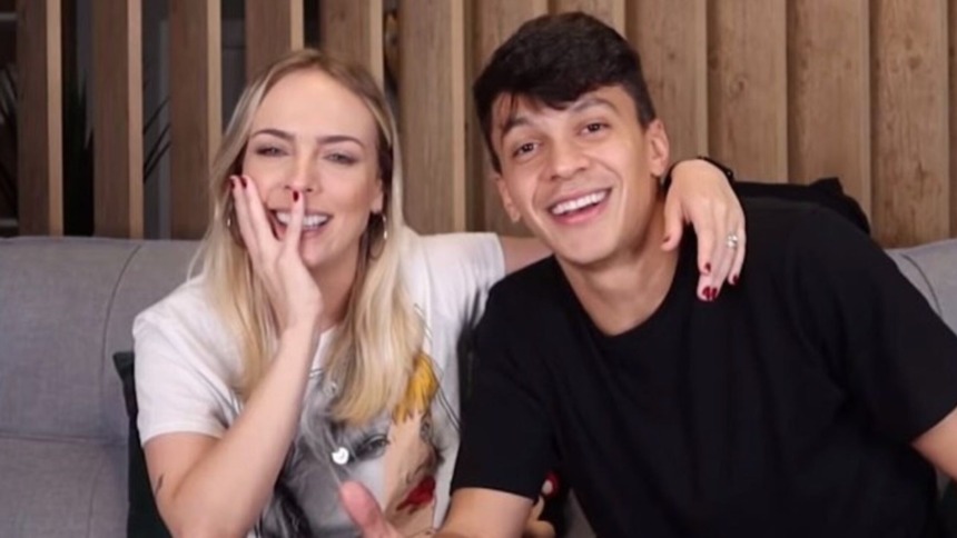 Os influenciadores e apresentadores Júlio Cocielo (camiseta preta) e Tata Estaniecki (cabelo loiro, camiseta branca) sorriem e se abraçam em um sofá.