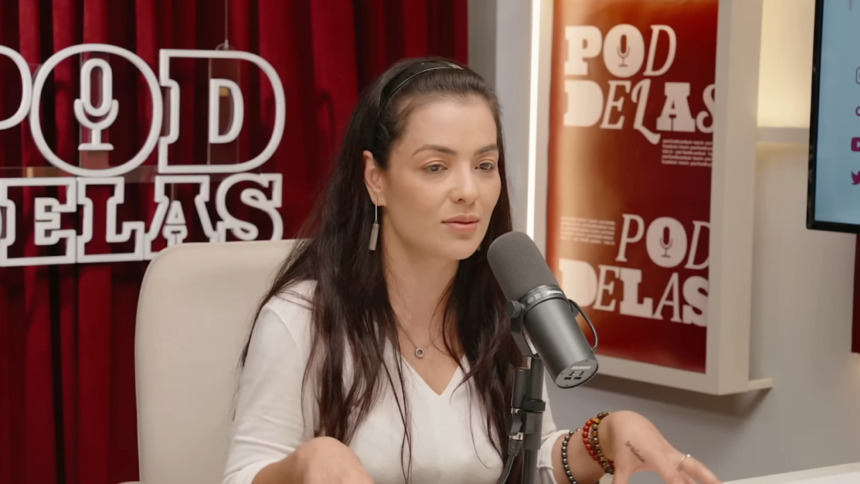 A cantora Li Martins (cabelos escuros e longos, blusa branca) está sentada em uma cadeira clara, falando em um microfone profissional durante uma entrevista no Pod Delas.