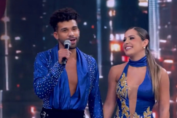 O ator Lucas Leto e a dançarina Mayara Araújo no palco do Dança dos Famosos. Ele está de camisa azul de cetim brilhante e aberta no peito, segurando um microfone. Ela usa um vestido azul com detalhes dourados no busto e está sorridente.