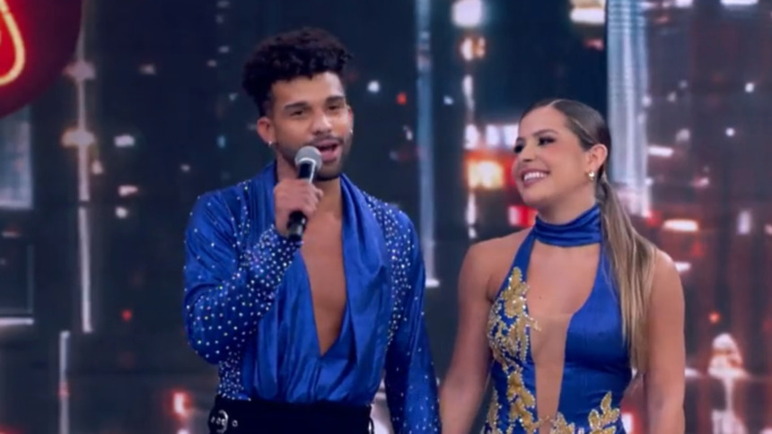 O ator Lucas Leto e a dançarina Mayara Araújo no palco do Dança dos Famosos. Ele está de camisa azul de cetim brilhante e aberta no peito, segurando um microfone. Ela usa um vestido azul com detalhes dourados no busto e está sorridente.