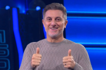 O apresentador Luciano Huck sorri e faz "joinha" com as duas mãos no palco azul do programa The Wall. Ele usa um suéter cinza.
