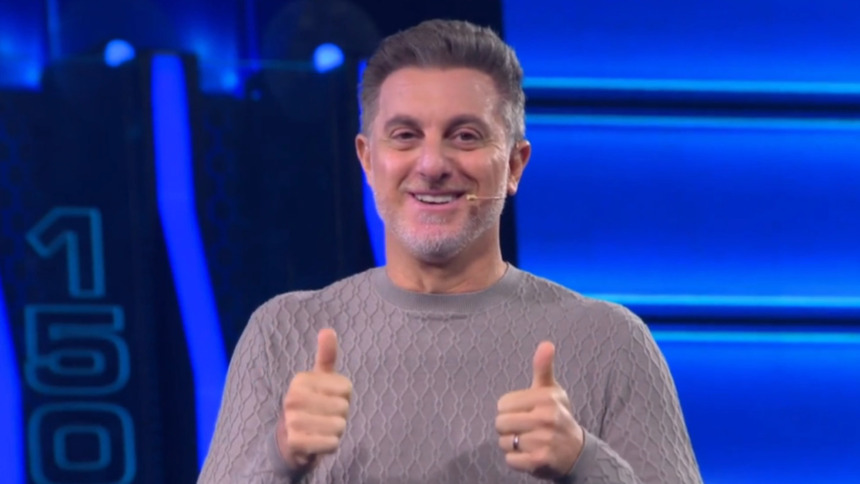 O apresentador Luciano Huck sorri e faz "joinha" com as duas mãos no palco azul do programa The Wall. Ele usa um suéter cinza.