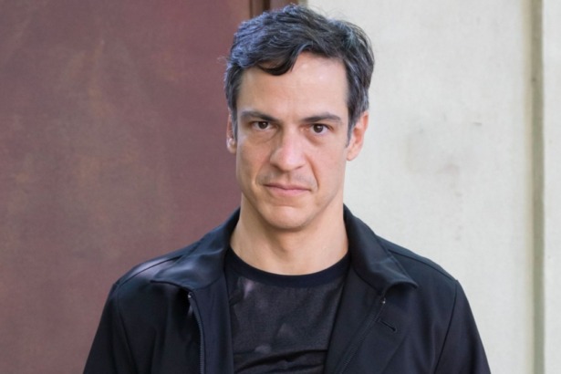 Retrato do ator Mateus Solano. Ele tem cabelos escuros e veste uma jaqueta preta sobre uma camiseta escura, olhando com uma expressão séria.