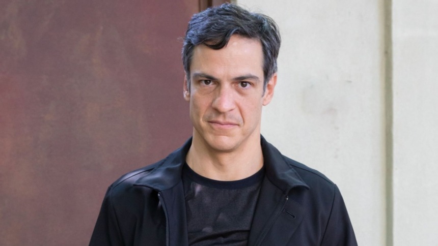 Retrato do ator Mateus Solano. Ele tem cabelos escuros e veste uma jaqueta preta sobre uma camiseta escura, olhando com uma expressão séria.