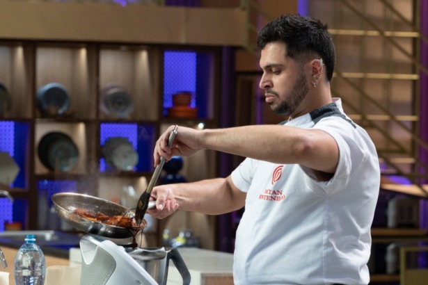 O cozinheiro Natan Montenegro no MasterChef Confeitaria está na cozinha, vestido com dólmã branco, concentrado e usando uma pinça para manusear alimentos em uma pequena frigideira.