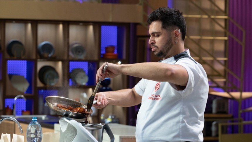 O cozinheiro Natan Montenegro no MasterChef Confeitaria está na cozinha, vestido com dólmã branco, concentrado e usando uma pinça para manusear alimentos em uma pequena frigideira.