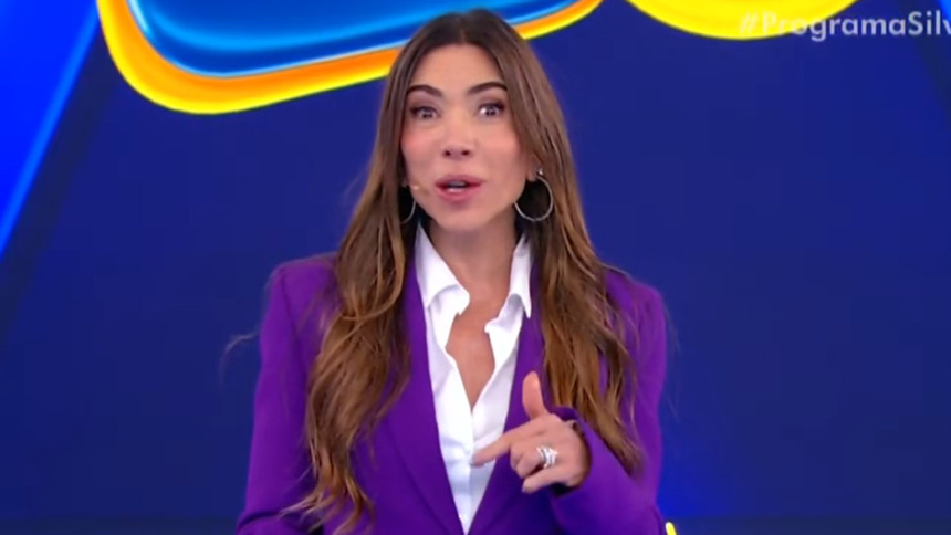 A apresentadora Patricia Abravanel no palco, de blazer roxo sobre camisa branca, sorri e aponta. O fundo é azul, da Tele Sena de Primavera.