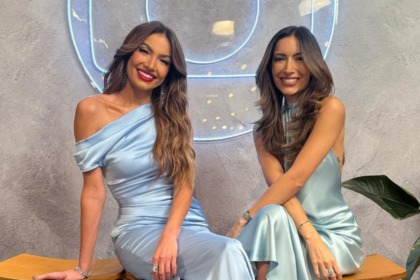 As apresentadoras Patrícia Poeta (vestido cetim azul-bebê de um ombro só) e Paloma Poeta (vestido cetim azul-bebê com gola alta) estão sentadas e sorrindo, em um ensaio fotográfico para a vinheta de fim de ano da Globo.