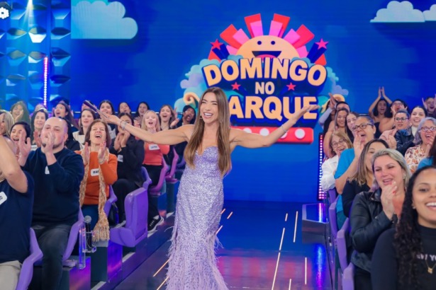 Patricia Abravanel usa um vestido roxo de paetês no palco do Domingo no Parque. Ela sorri, de braços abertos, em frente à plateia do Programa Silvio Santos.