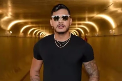 O rapper Hungria está em um túnel dourado. Ele usa camiseta preta, óculos escuros e colares de corrente prateada, exibindo tatuagens nos braços.