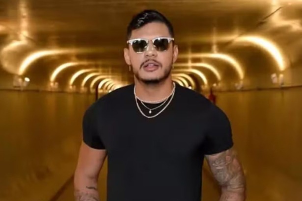 O rapper Hungria está em um túnel dourado. Ele usa camiseta preta, óculos escuros e colares de corrente prateada, exibindo tatuagens nos braços.