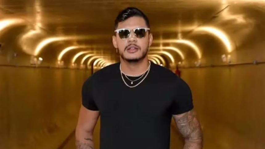 O rapper Hungria está em um túnel dourado. Ele usa camiseta preta, óculos escuros e colares de corrente prateada, exibindo tatuagens nos braços.