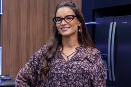 A chef Renata Vanzetto em um estúdio de cozinha do reality Chef de Alto Nível. Ela usa óculos de gatinho, uma blusa floral em tons escuros e sorri para a câmera.