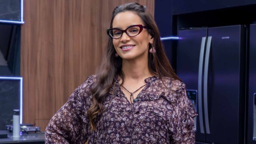 A chef Renata Vanzetto em um estúdio de cozinha do reality Chef de Alto Nível. Ela usa óculos de gatinho, uma blusa floral em tons escuros e sorri para a câmera.