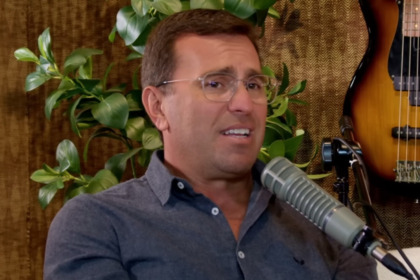 O jornalista Rodrigo Bocardi está em uma entrevista para um podcast falando sobre a Globo. Ele usa óculos de grau, camisa azul-maro escuro de botões e fala em um microfone profissional.