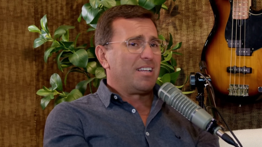 O jornalista Rodrigo Bocardi está em uma entrevista para um podcast falando sobre a Globo. Ele usa óculos de grau, camisa azul-maro escuro de botões e fala em um microfone profissional.