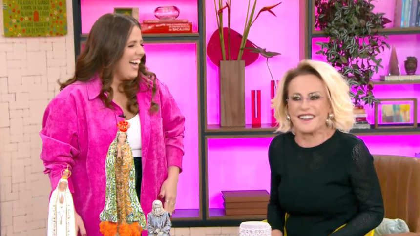 As apresentadoras Tati Machado (blazer rosa choque) e Ana Maria Braga (blusa preta com mangas furadas) estão sorridentes no cenário do Mais Você.