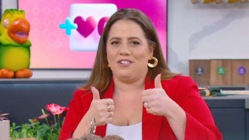 Tati Machado sorri e faz "joinha" com as duas mãos. Ela veste um blazer vermelho no cenário colorido do programa Mais Você, anunciando que vai divulgar Três Graças no intervalo de Vale Tudo