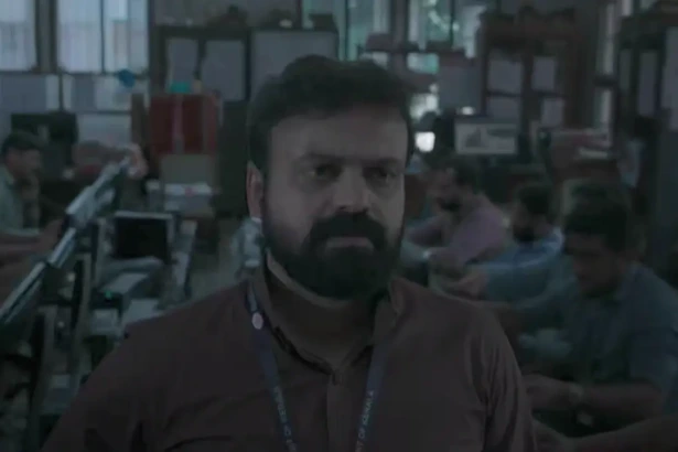 Homem com crachá no pescoço olha para um telão assustado numa sala de controle em cena do filme Todos Somos Heróis
